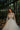 A woman wearing a white, floral lace wedding dress with a sweetheart neckline and sleeveless design, standing in a room with a reflective floor and greenery in the background.
